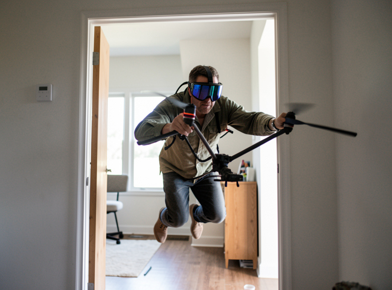 A pilot flying a cinewhoop drone indoors through a doorway while wearing FPV goggles